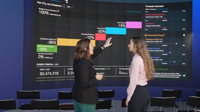 Two women stand if front of large digital screen displaying charts and data related to emissions and forecast scenarios, discussing the information. 