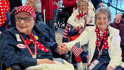 Two Rosie the Riveters hold hands, wearing red polka dot scarves and holding American flags.