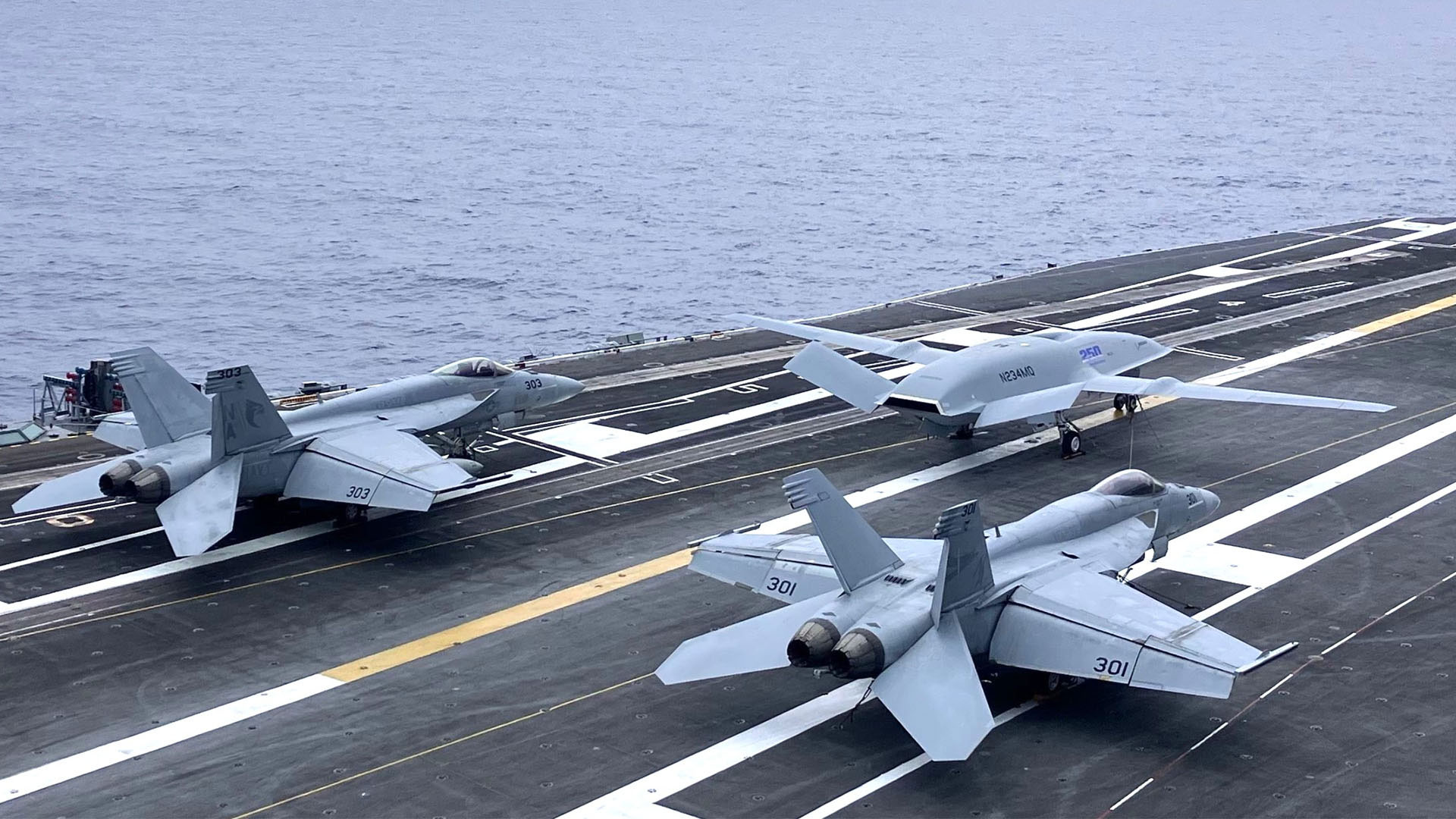 Boeing’s MQ-25A T1 prototype positioned alongside F/A-18 Super Hornets aboard the USS Nimitz. The aircraft features a special “Boeing Backs America” mark highlighting Boeing’s support for U.S. aviation.
