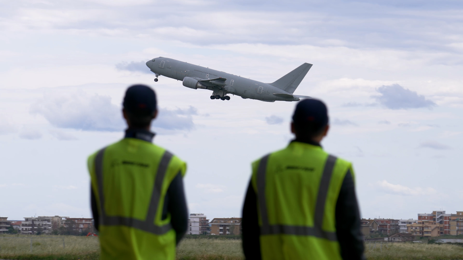 Two Boeing employees in bright yellow safety vests have their back to the camera as they watch an aircraft take off in cloudy skies.