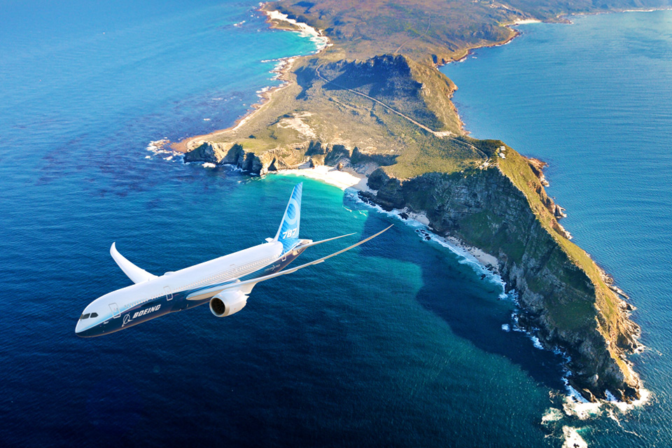 A Boeing 787 Dreamliner soaring over a scenic coastline, with lush greenery and blue waters below.