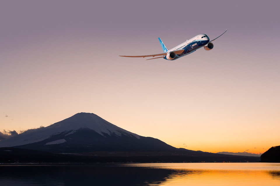 787 Dreamliner flying over a body of water with a mountain in the background.