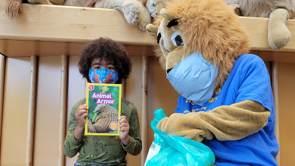 Child holding up book, beside lion mascot