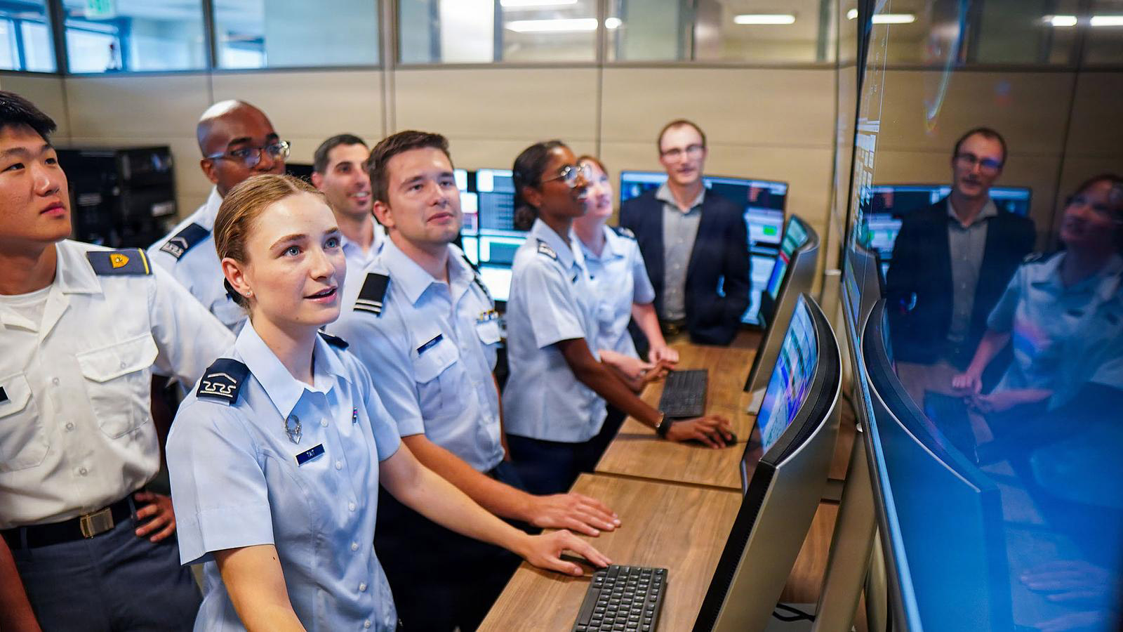 Female cadet smiling while looking at computer monitor. Several more cadets surrounding her. 