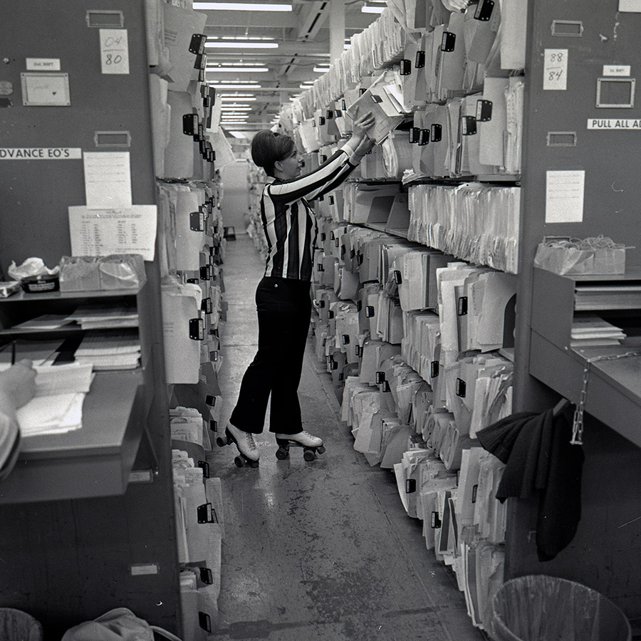 Black and white photo of woman in striped shirt putting files on shelf.