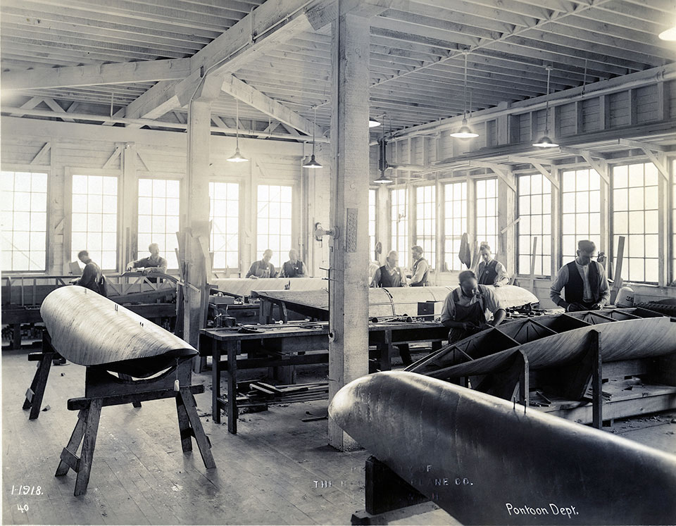 In 1918, Dick and George Pocock were crafting airplane pontoons in the Red Barn. Dick Pocock, wearing an open vest, is far right. George Pocock is working on the pontoon behind Dick, wearing coveralls, a vest and a necktie