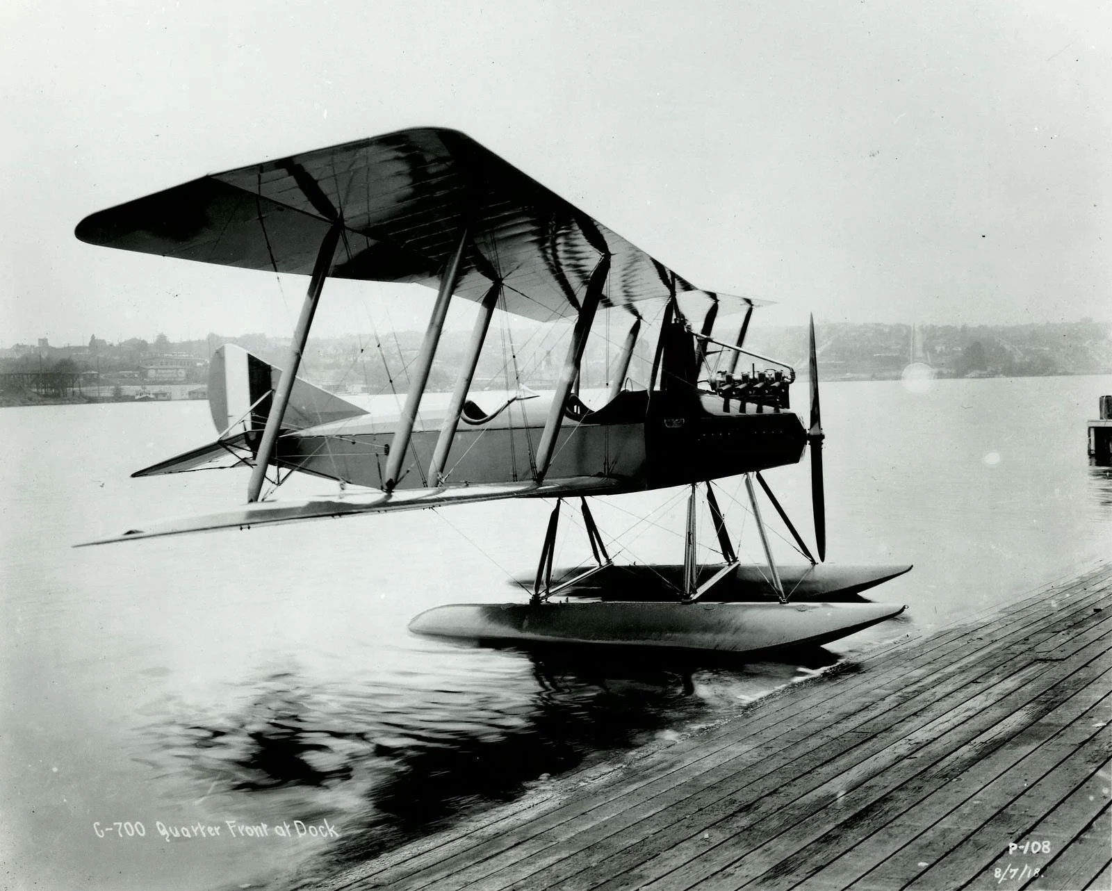 A black and white photo of the Model C, a biplane seaplane, sitting in the water at a dock.