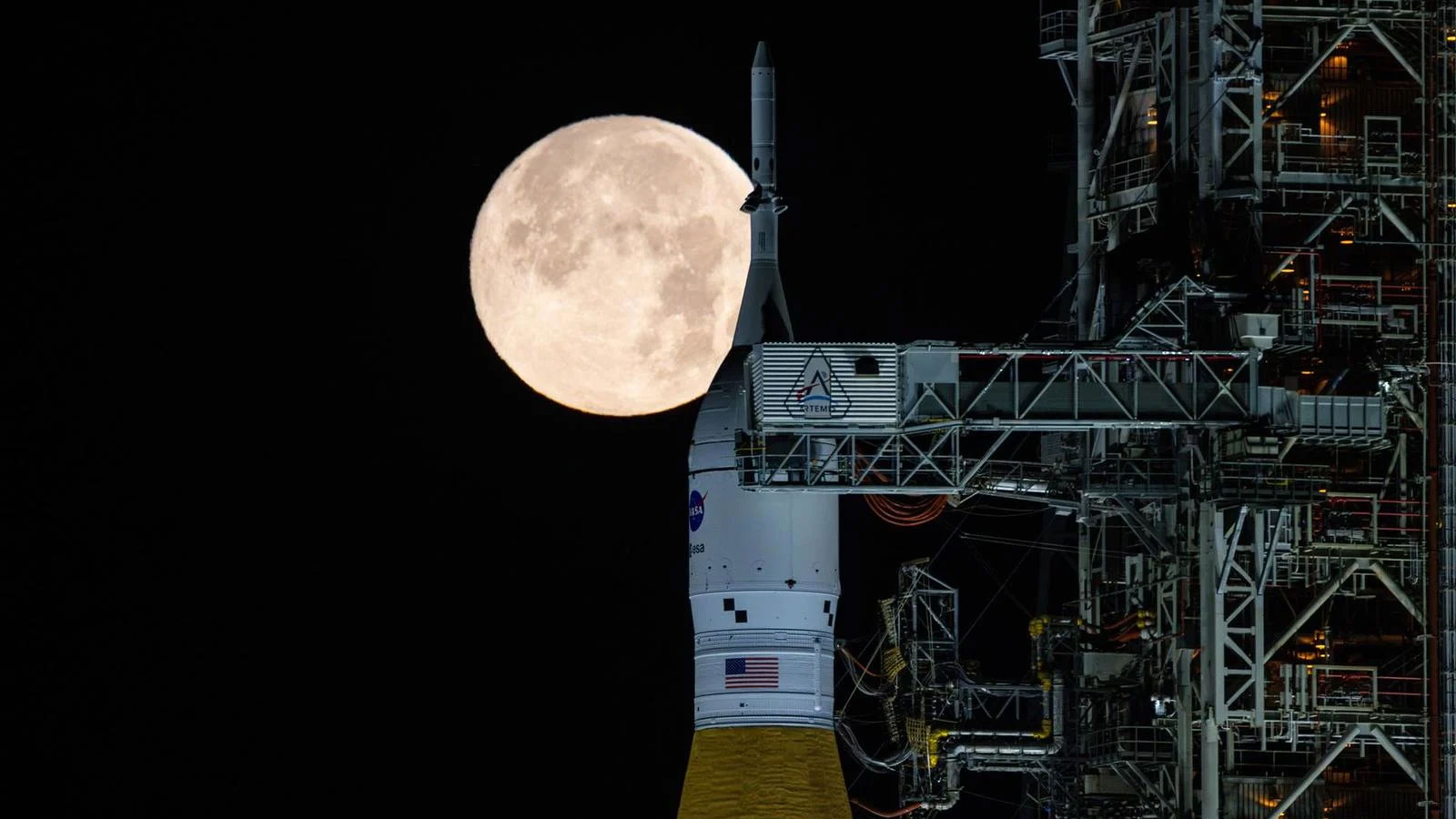 The Artemis II rocket sitting on the launch pad with a full moon behind it. 