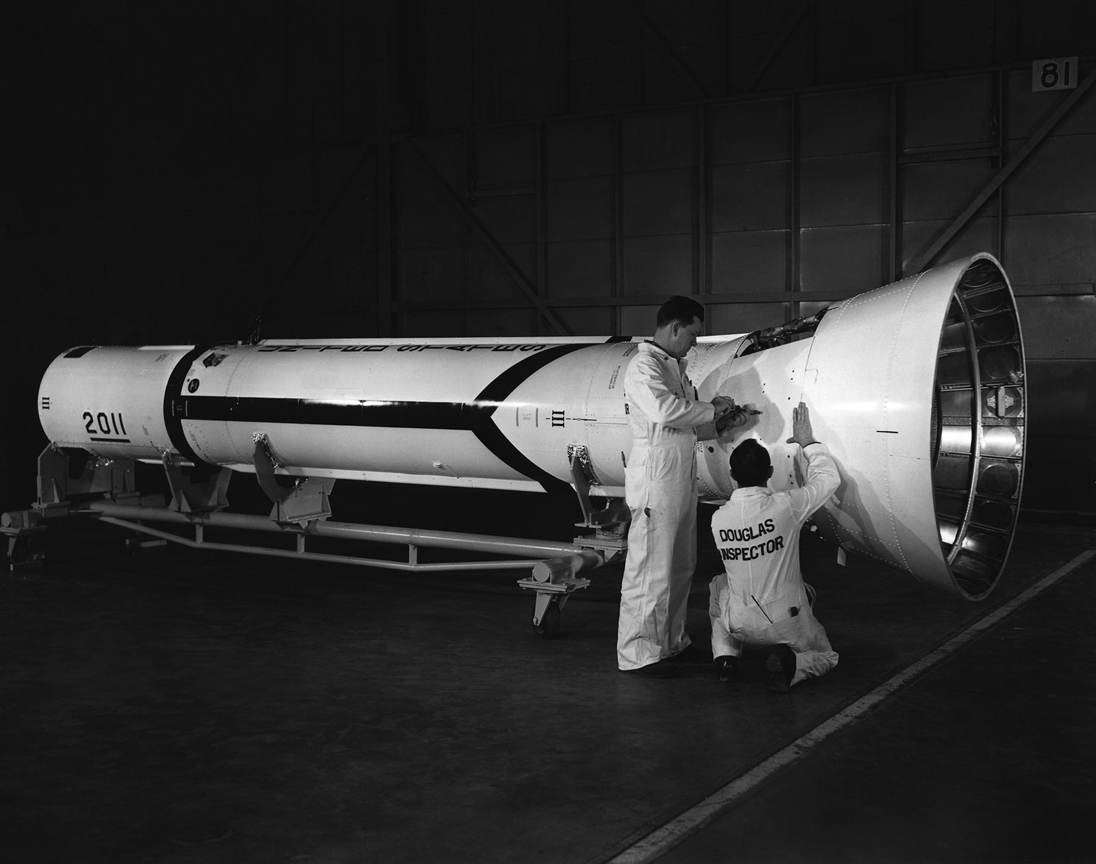 Black and white photo of two Douglas Inspectors looking at a horizontally-laying piece of a Delta rocket