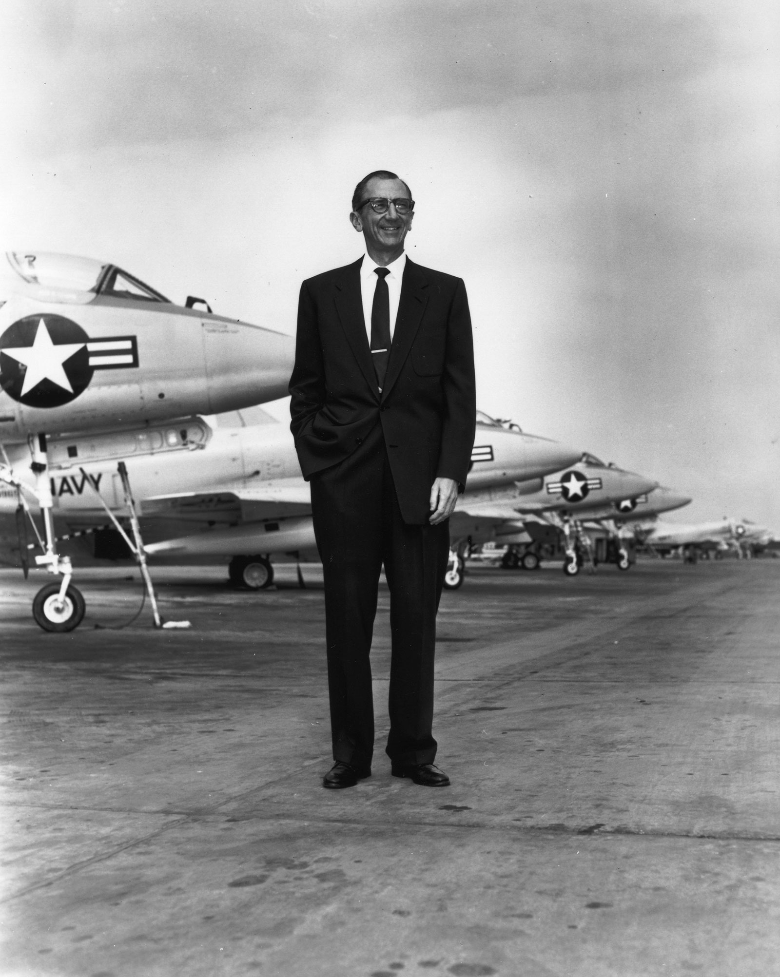 Black and white photo of Ed Heinemann, dressed in a black suit, in front of a row of A-4 Skyhawk jets.