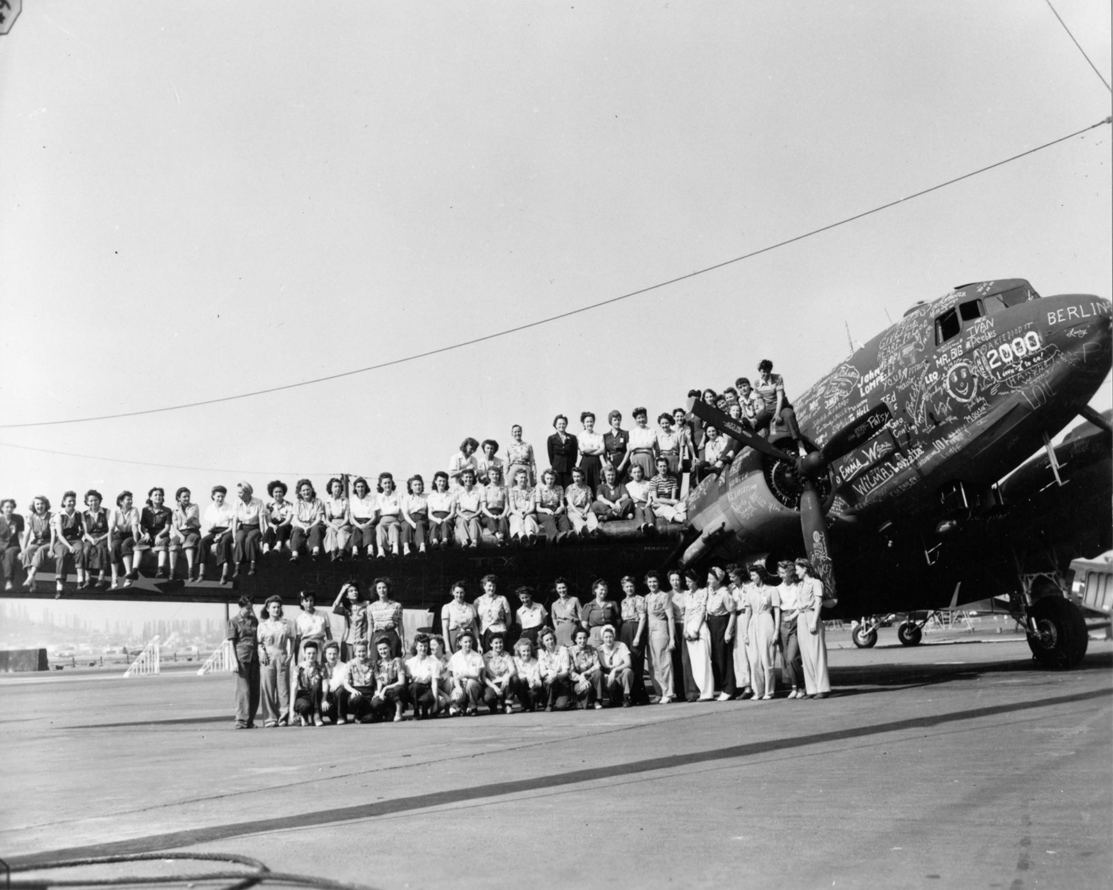 Black and white photo of a C-47, a two-engine propellor plane, covered in signatures of the workers who built it. Sitting on the wing and standing in front of the plane are many female mechanics who helped build it.