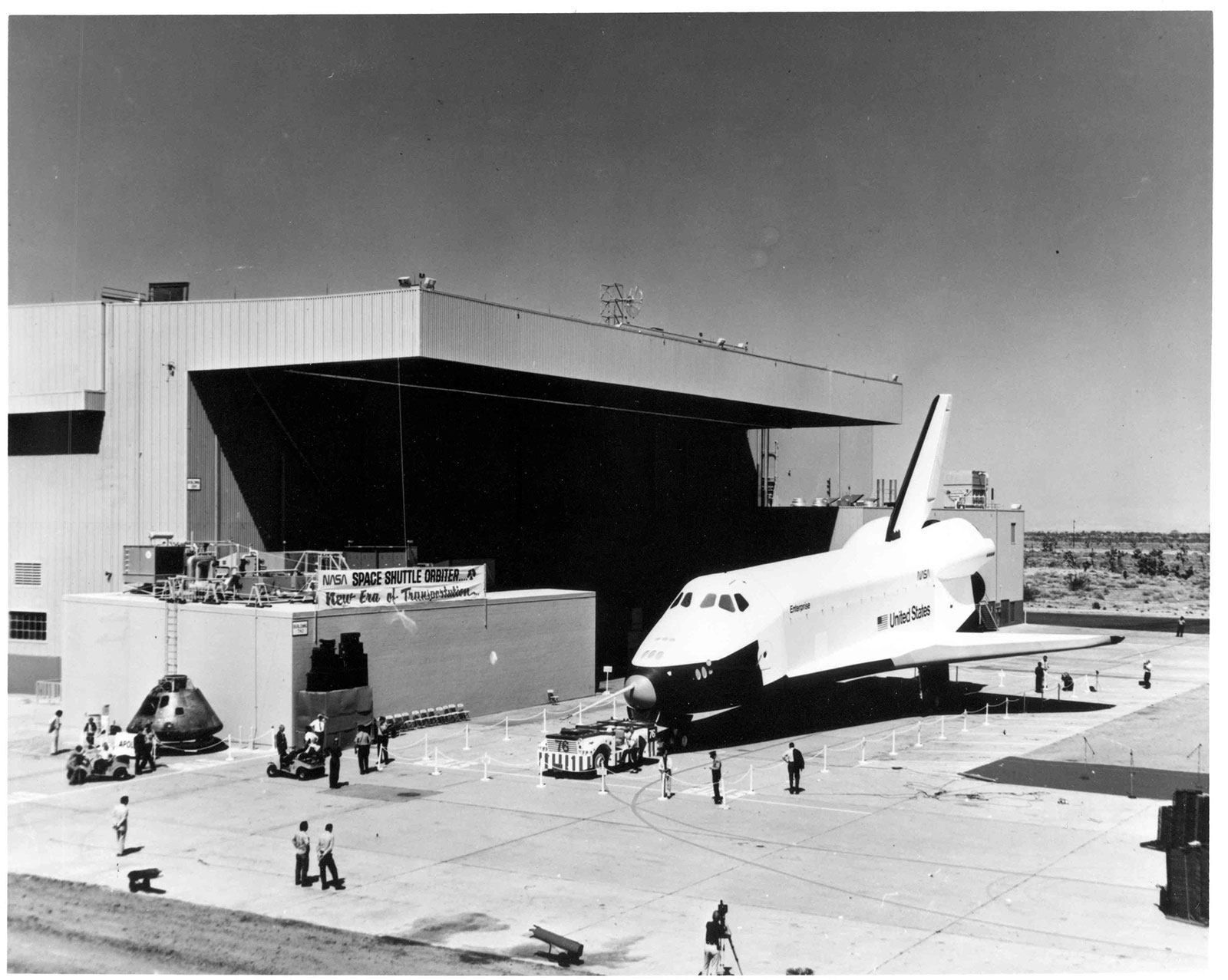 Black and white photo of the Space Shuttle Orbiter sitting in front of a factory building. Nearby on the ground are several people and an Apollo Command Module. A large banner reads "NASA Space Shuttle Orbiter... New Era of Transportation..."