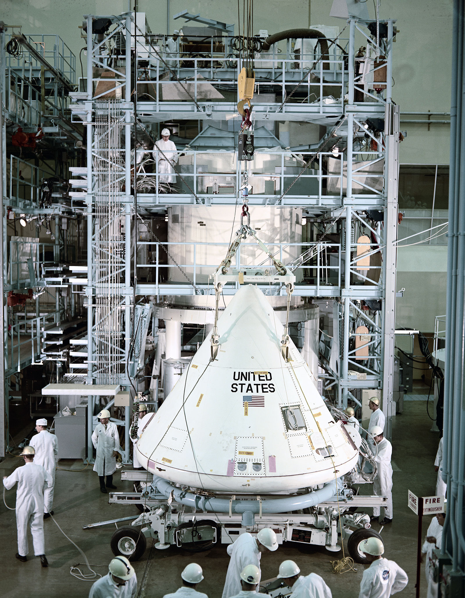 Color photo of the Apollo Command Module, painted white, in the factory with many workers around it