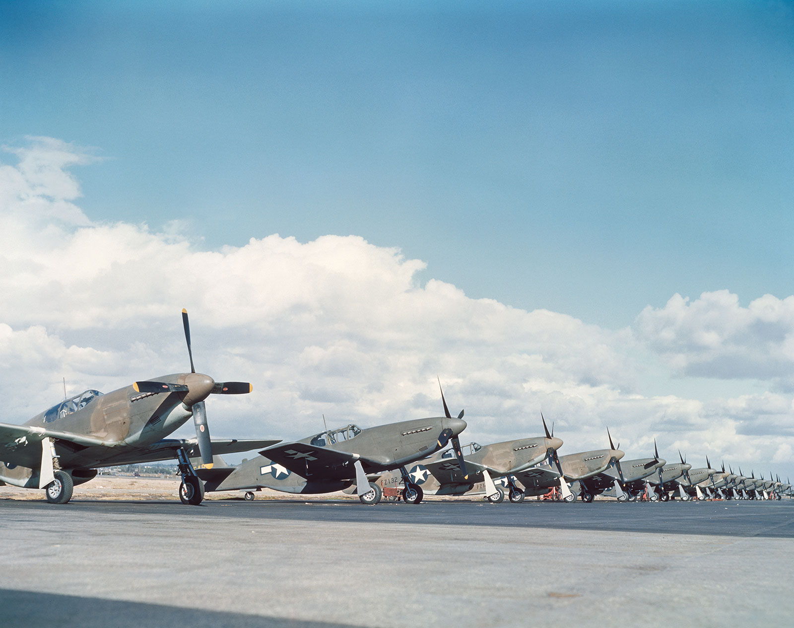 Color photo of dozens of P-51B Mustangs lined up on the tarmac against a blue sky