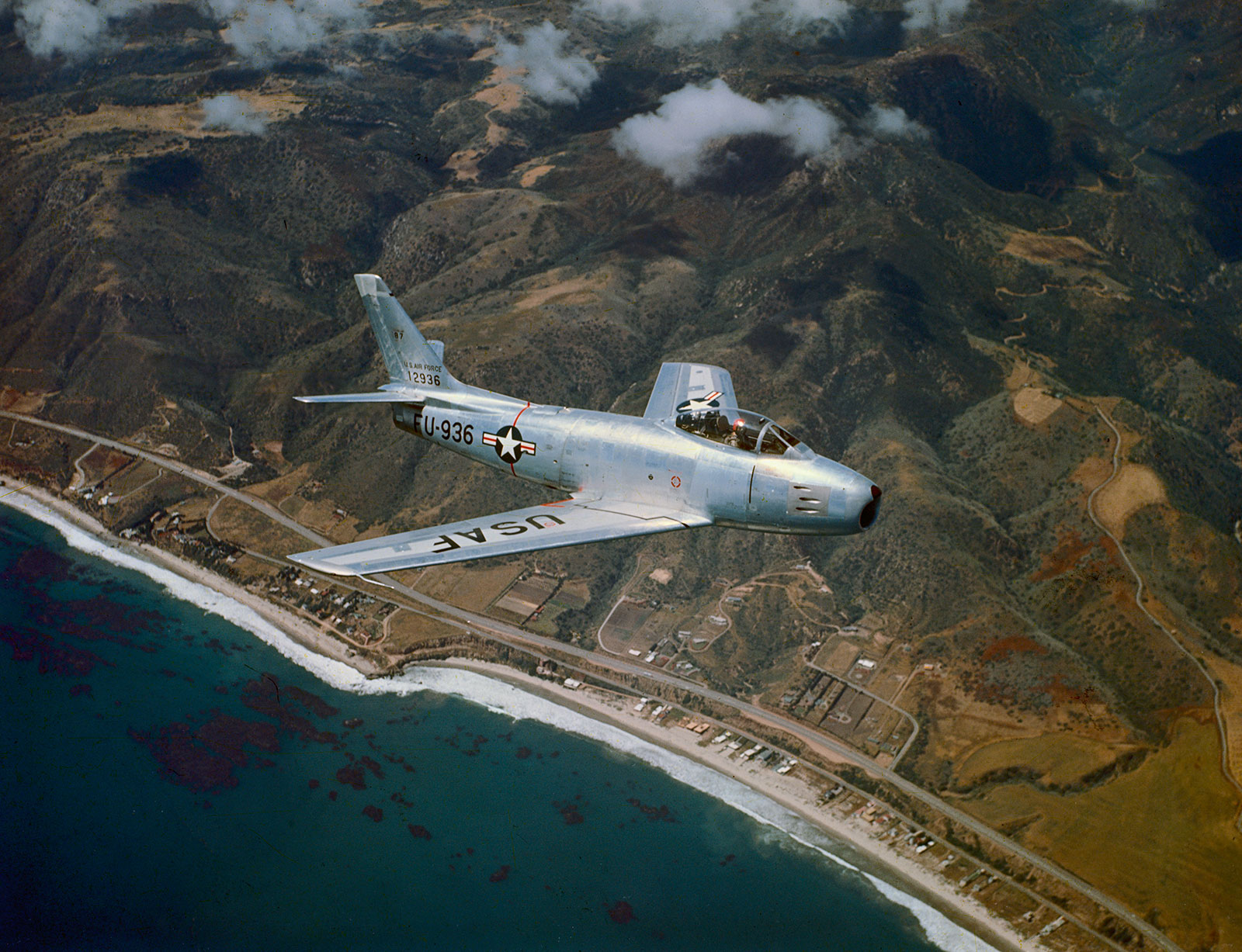 Color photo of a F-86 Sabre, a single jet engine fighter, in flight over a shoreline