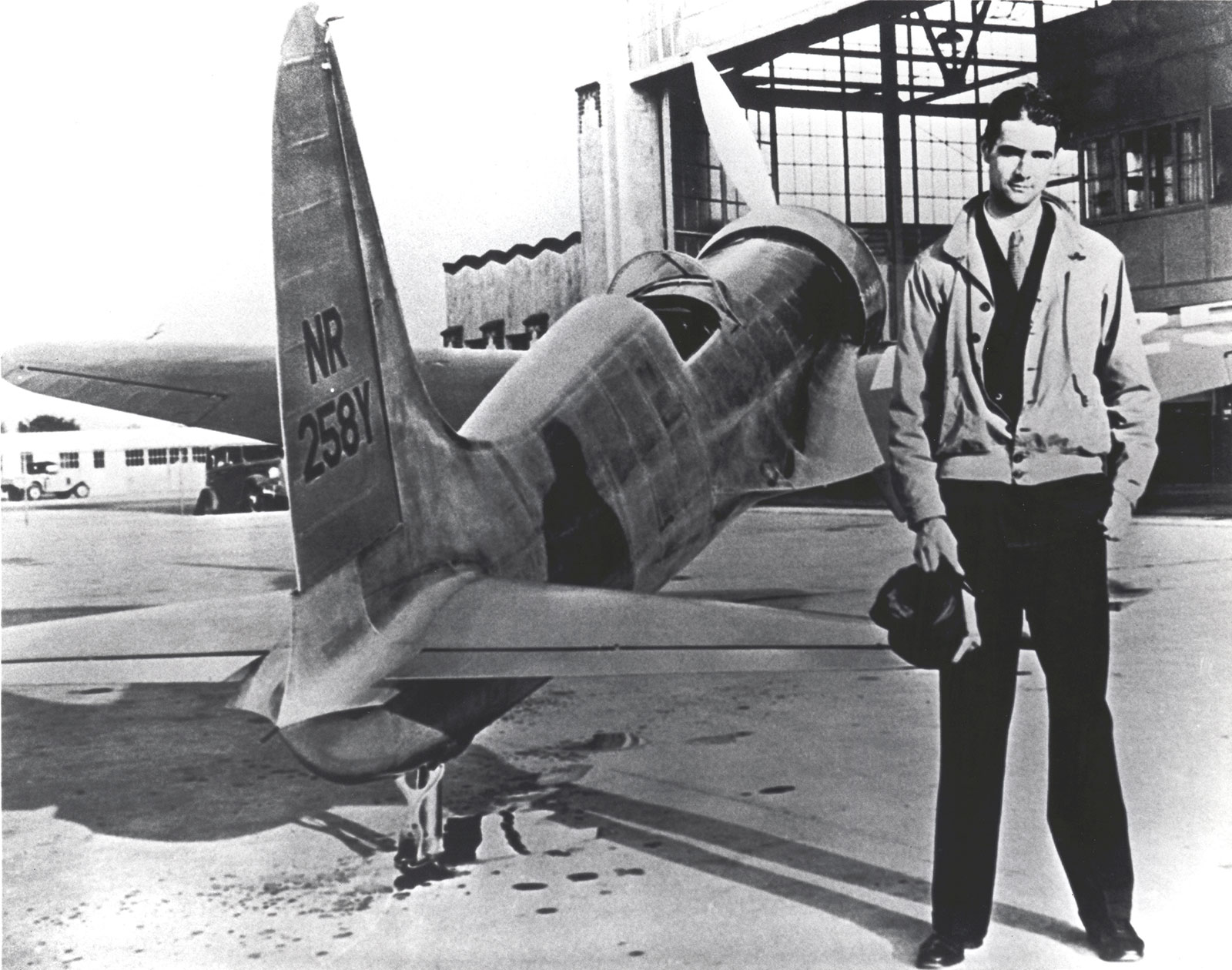 Black and white photo of Howard Hughes, wearing a flight jacket and holding his flight cap, next to the H-1 Racer. Hughes is standing by the tail of the aircraft.