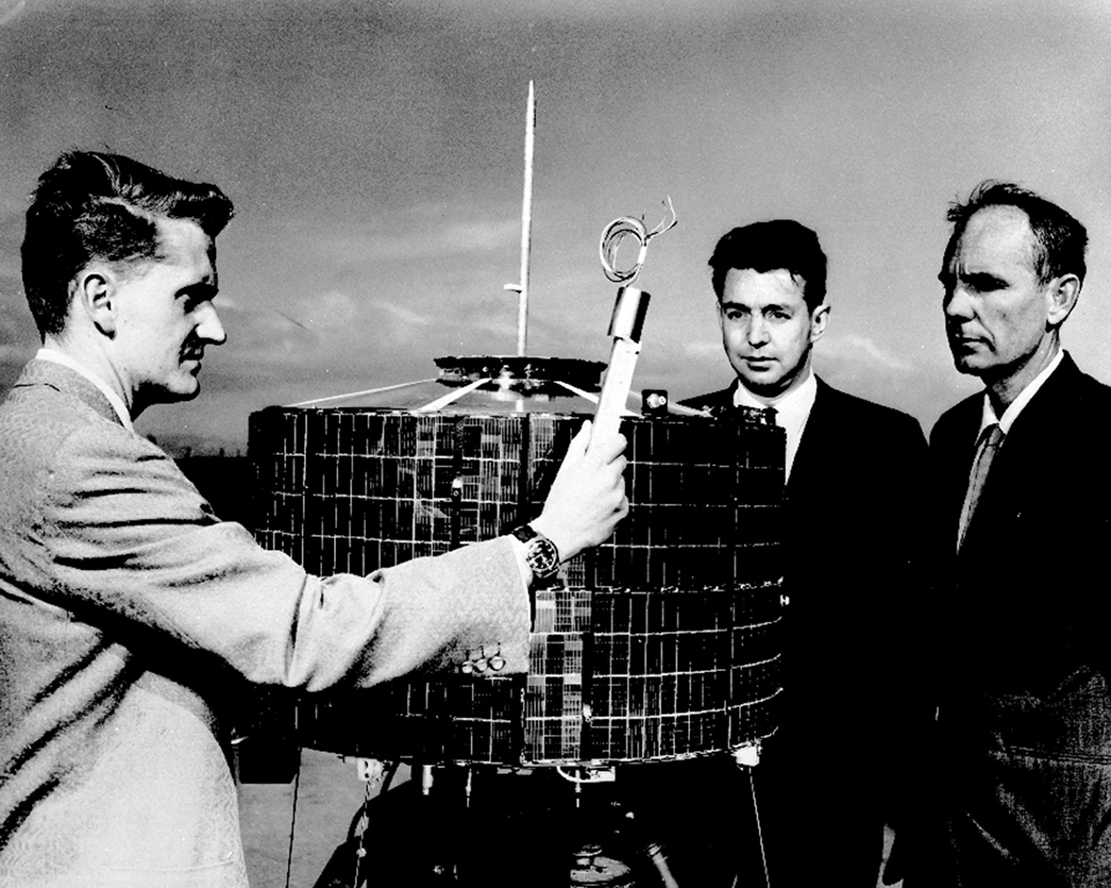 Black and white photo of Don Williams, Harold Rosen, and Thomas Hudspeth standing around the Syncom satellite. Williams holds a piece of equipment in his right hand.