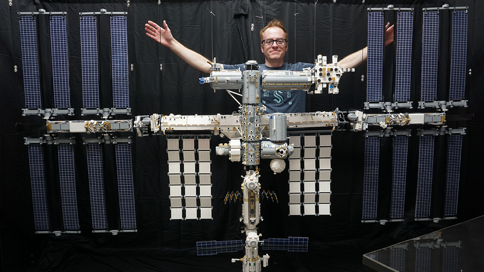 Doug Hughes with arms outstretched stands behind an 8-foot-wide, 1:42-scale LEGO model of the International Space Station.