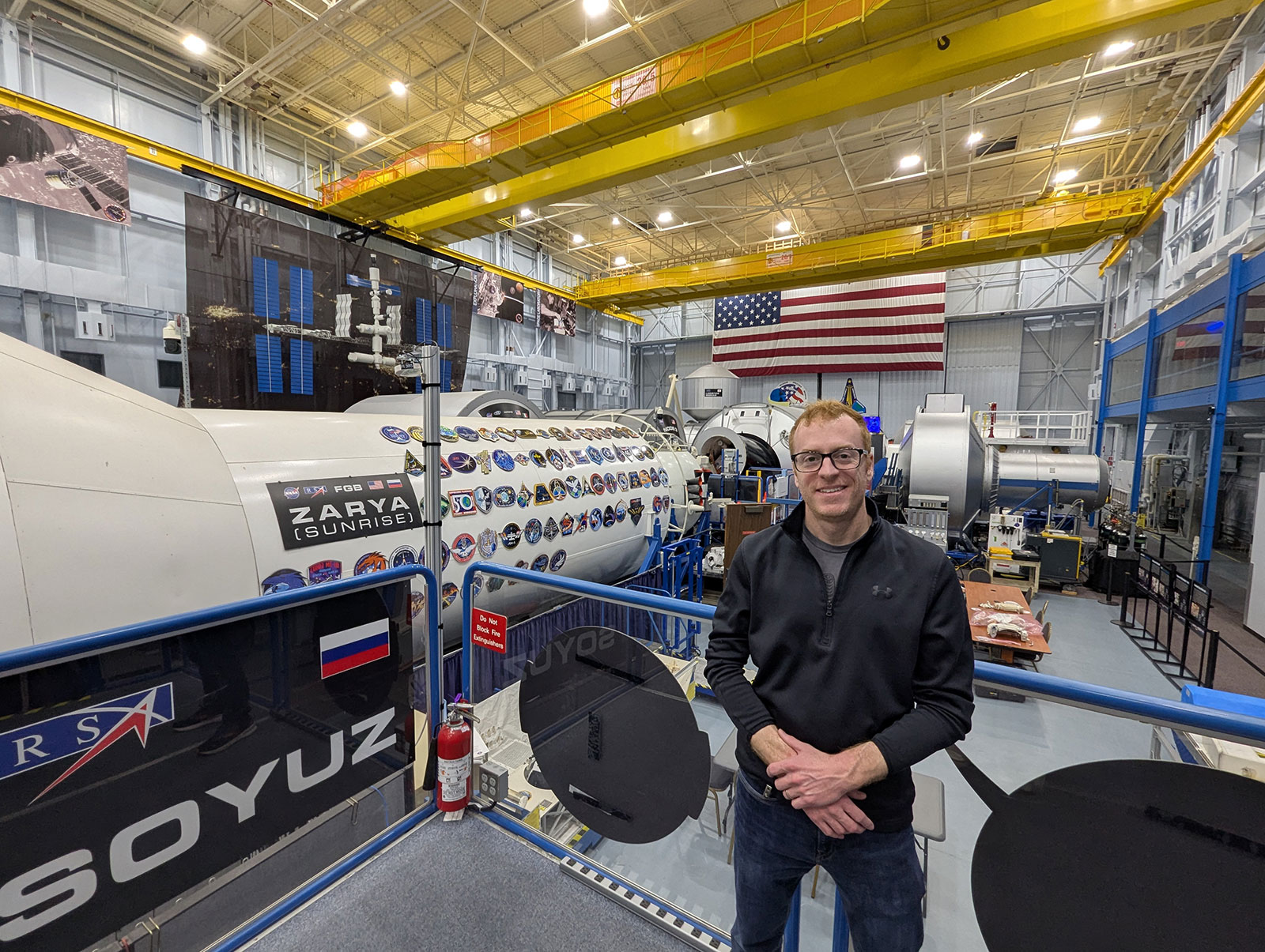 Hughes stands in a tour area inside a large hangar-like mockup facility with space modules and a U.S. flag.