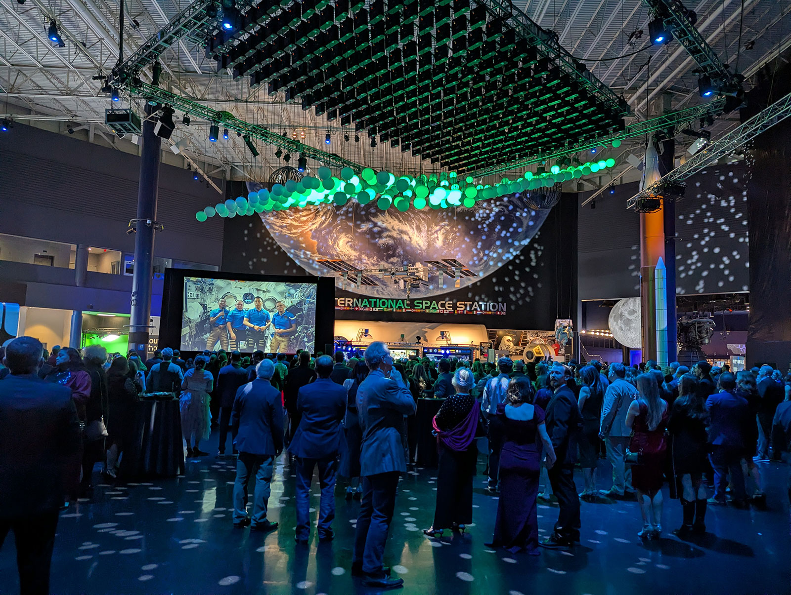 Crowd gathered in a dim museum hall facing a stage and large Earth backdrop under a ceiling installation of green lights.