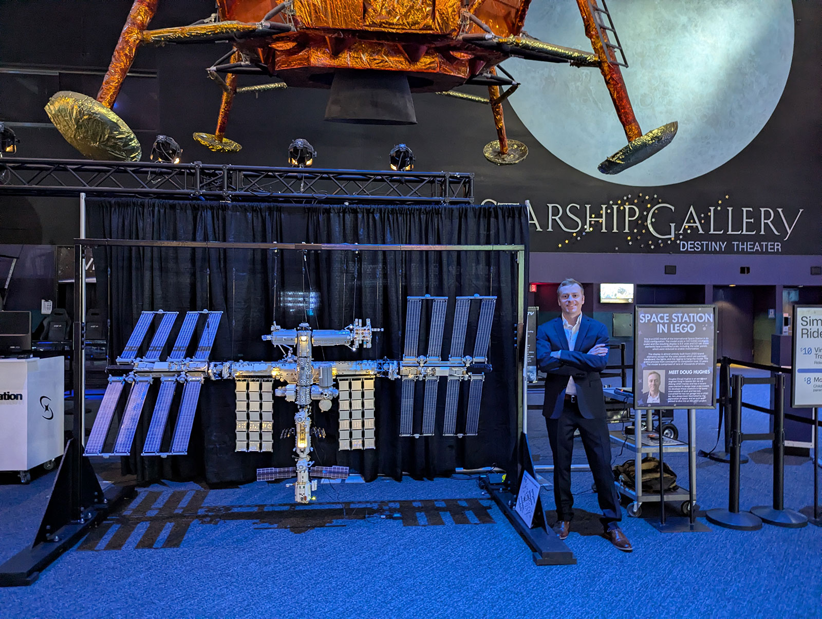 Hughes' standing beside a large LEGO International Space Station model on display in a museum gallery with a lunar mural behind them.