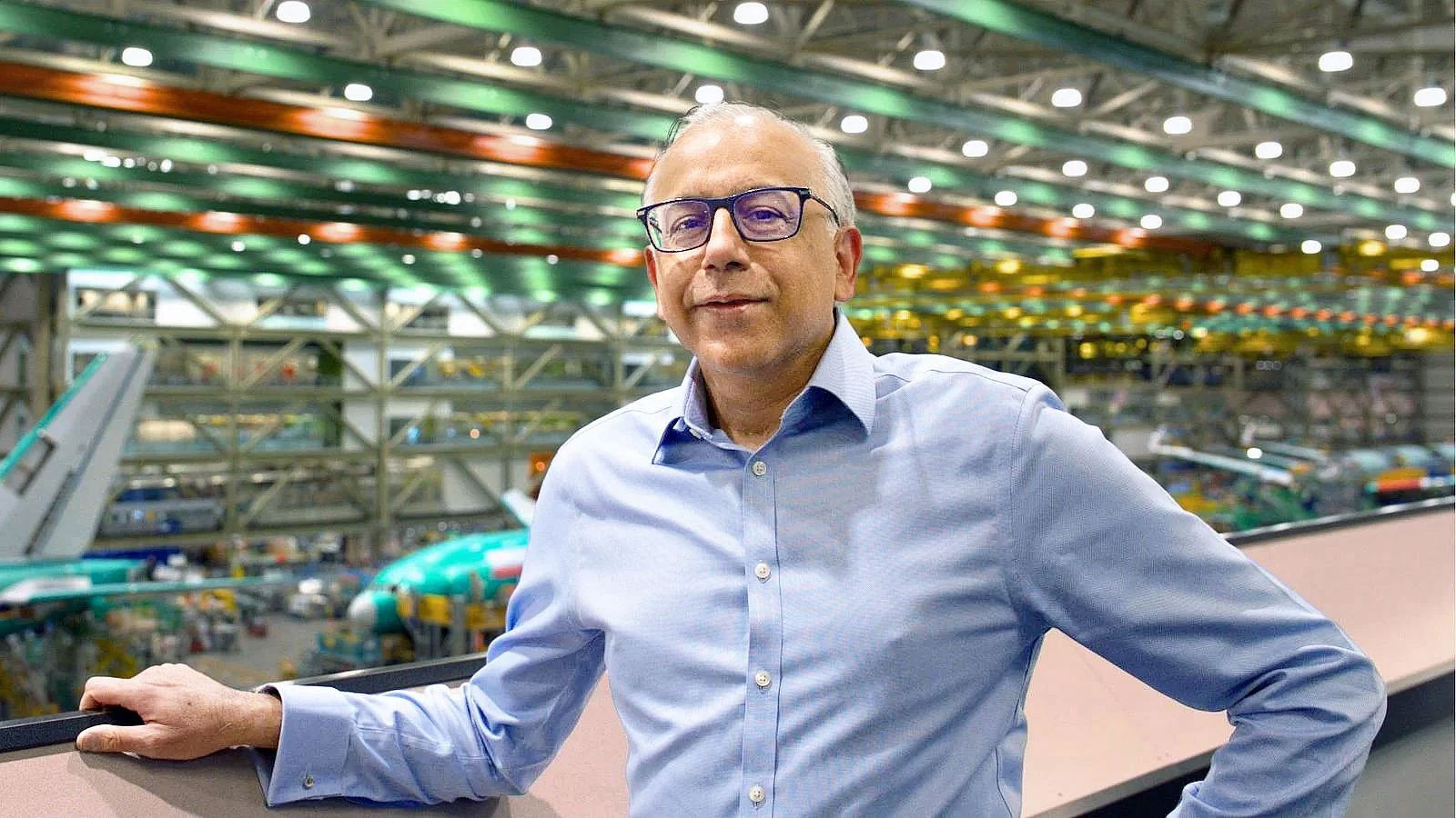 Portrait-format photo of Ratan Khatwa, an engineer, standing in the Boeing factory with blurred airplanes in the background; he is looking toward the camera and wearing business-casual attire. (Maddie Crisalli photo © Boeing) 