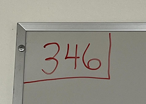 Close-up of the top-left corner of a whiteboard framed in aluminum; the large red marker digits '346' are written across the board and underlined with a red horizontal stroke.