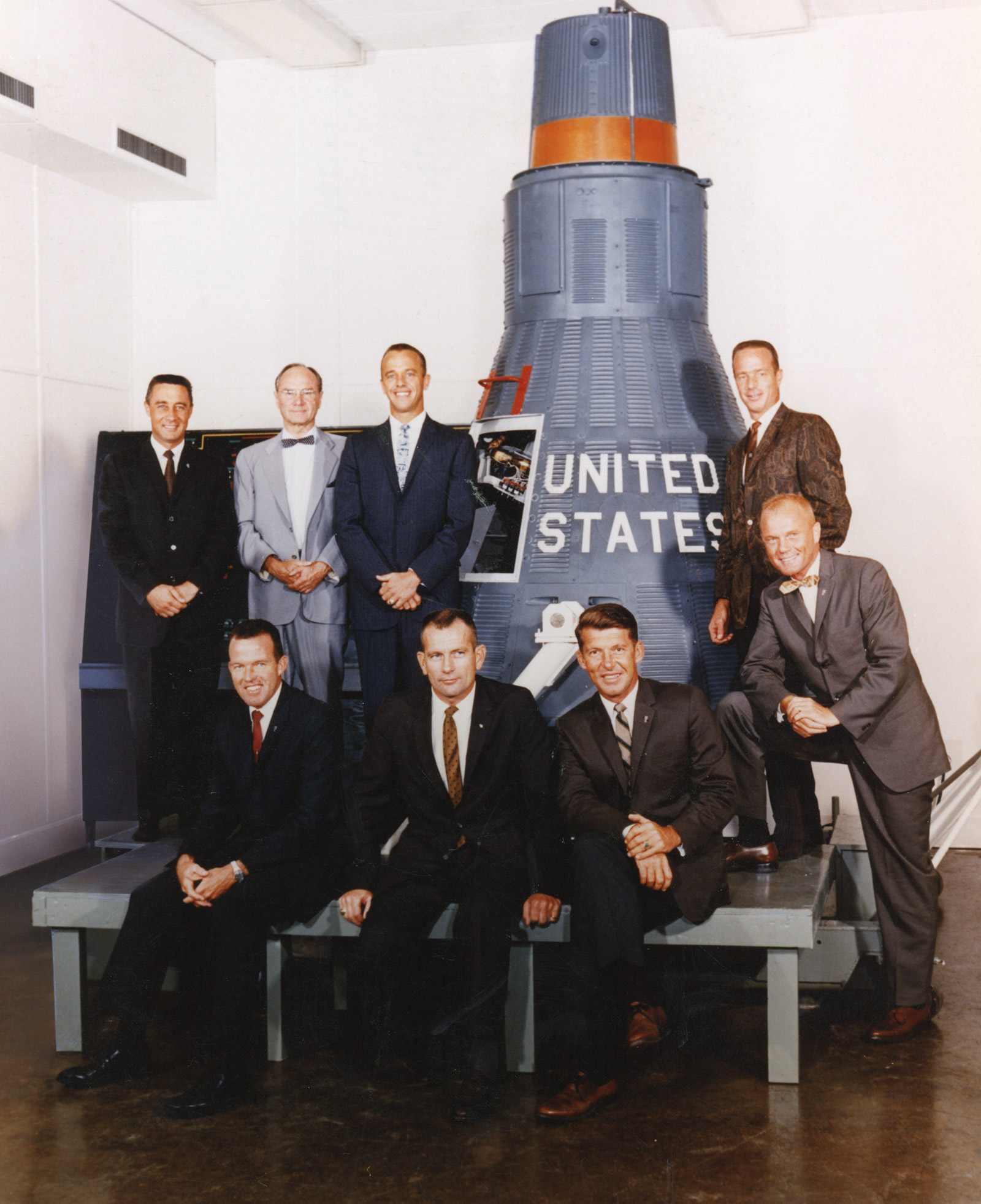 Color photo of seven men in suits posing for a photo in front of early space craft