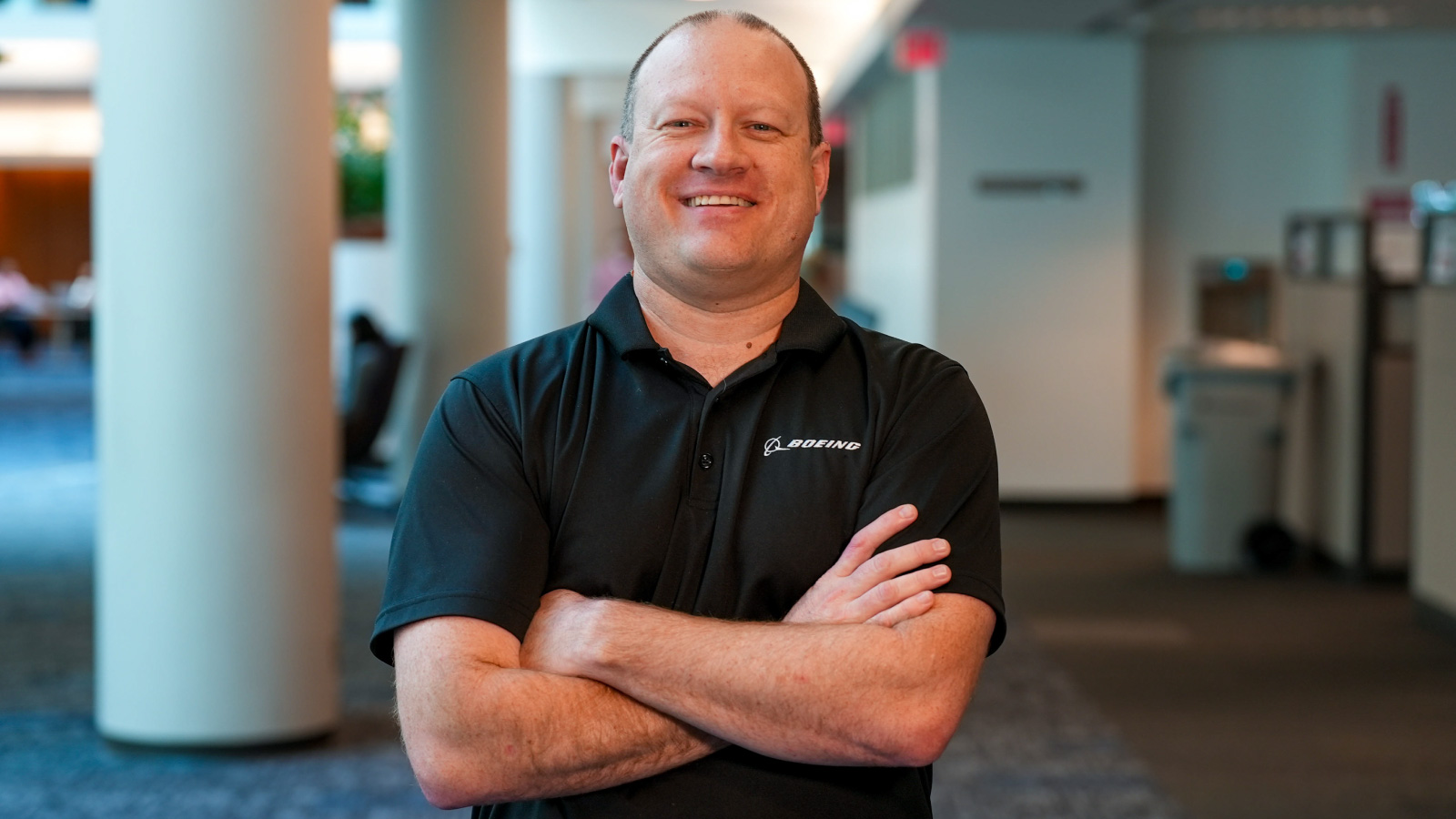 Flight‑sciences engineer standing in an office lobby with arms crossed, wearing a black polo with a company logo; columns and carpeted floor visible in the background.