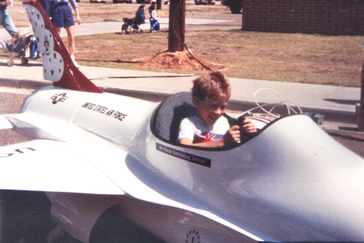 Child seated inside a small replica jet ride at an outdoor fair, gripping the controls; spectators and a tree are visible in the background.