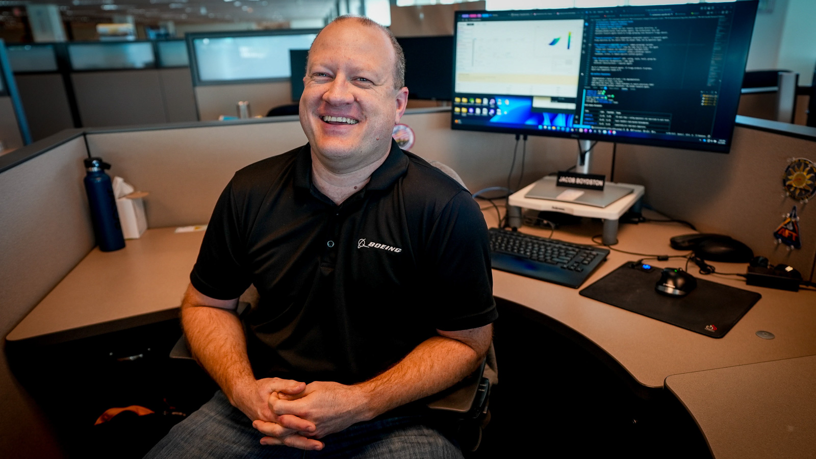 Engineer seated at a cubicle desk in front of dual monitors and keyboard, hands clasped, wearing a black polo; water bottle and desk items at the sides.