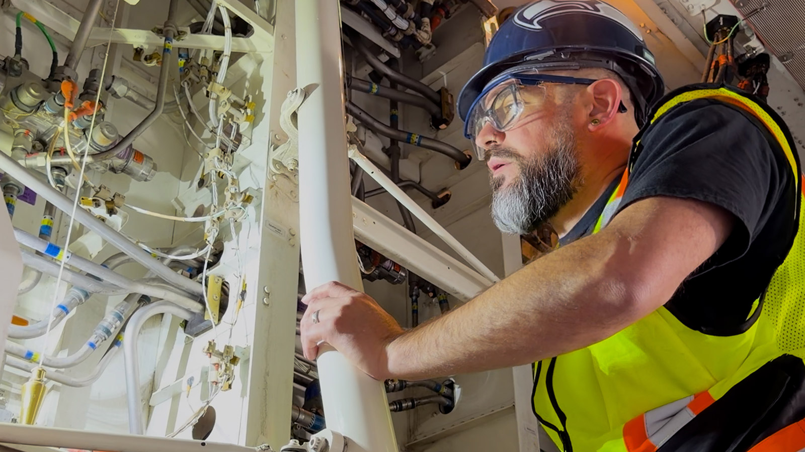 Will Santeo inspects a 787 Dreamliner. He is on a 10-foot-high ladder in the belly of the aircraft. Santeo is wearing a navy blue bump cap, reflective vest over a black t-shirt, and clear safety glasses with a blue frame.. He is using a fixed light to see the wiring inside the airplane.