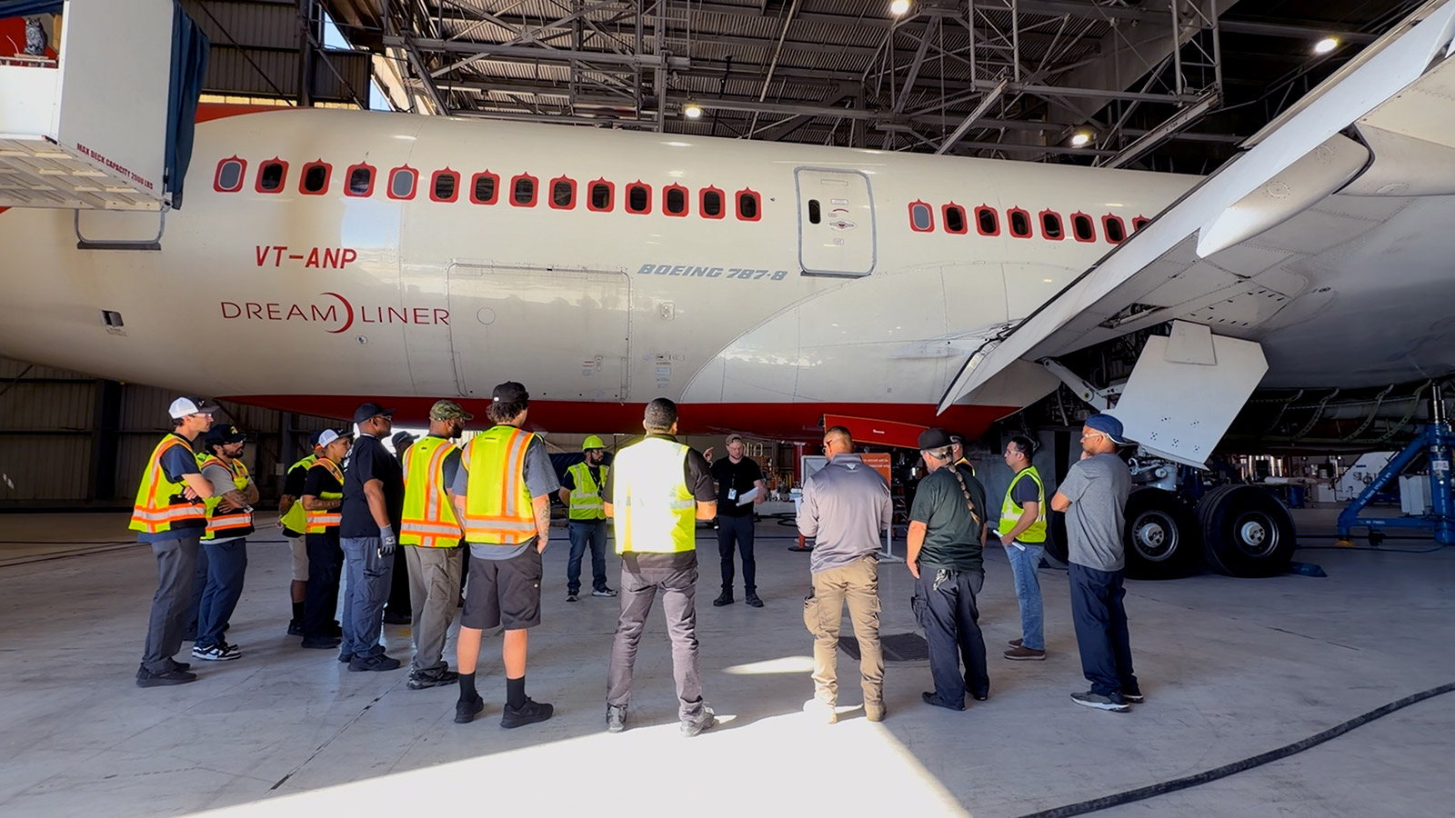 A team of at least 16 modification technicians stand in a circle to the right of the starboard side of a 787 Dreamliner, which is parked inside a hangar at the Global Services Modification Center in Victorville, California. They are reviewing their responsibilities for the day before they start working on the airplane. The manager, wearing a black shirt and backwards light brown cap, is reading a sheet of paper with each employee's job description to the team.