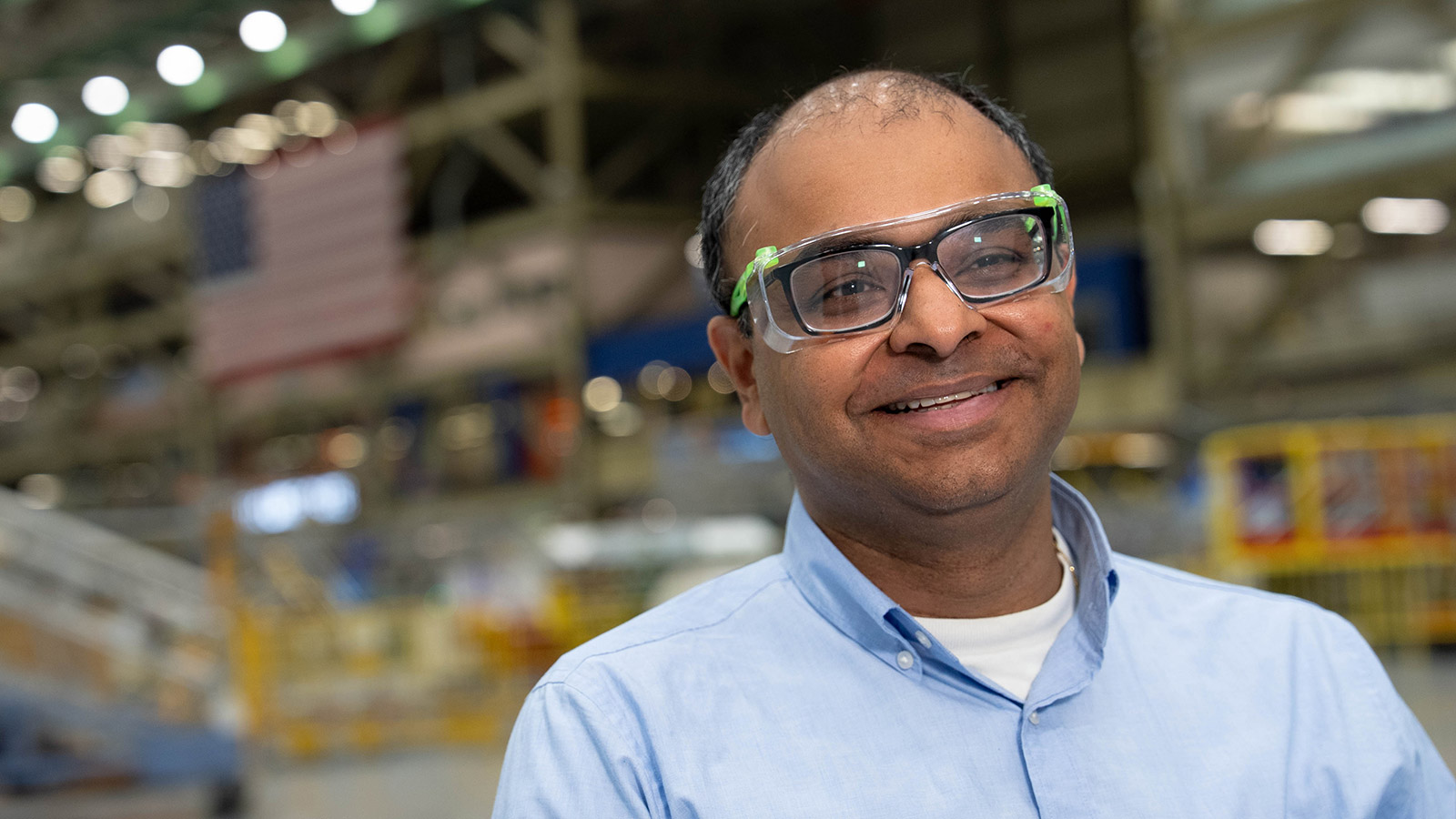 A medium-close portrait of a smiling man wearing clear safety glasses and a light blue button-up shirt, standing inside a large industrial hangar. The background is softly out of focus and shows bright overhead lights, structural beams, and a blurred American flag.