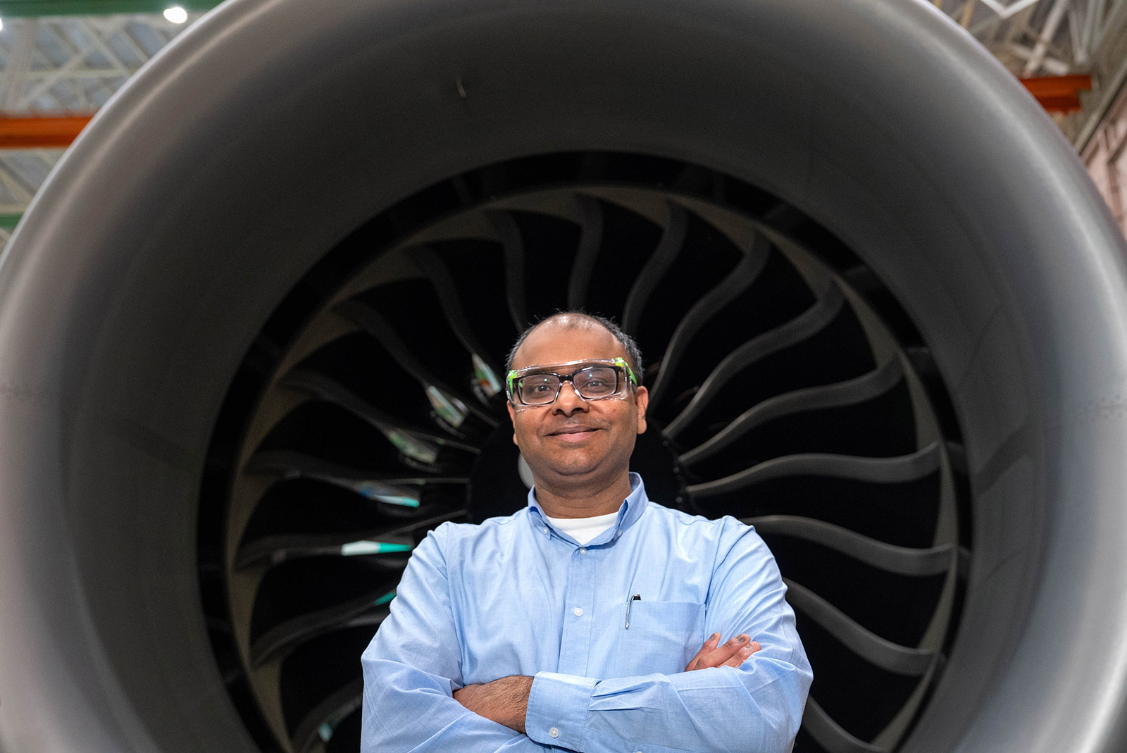 A wider shot of the same man standing with his arms crossed in front of a large jet engine. He wears safety glasses and a light blue shirt; the dark engine fan blades form a circular, ribbed backdrop that fills most of the frame.