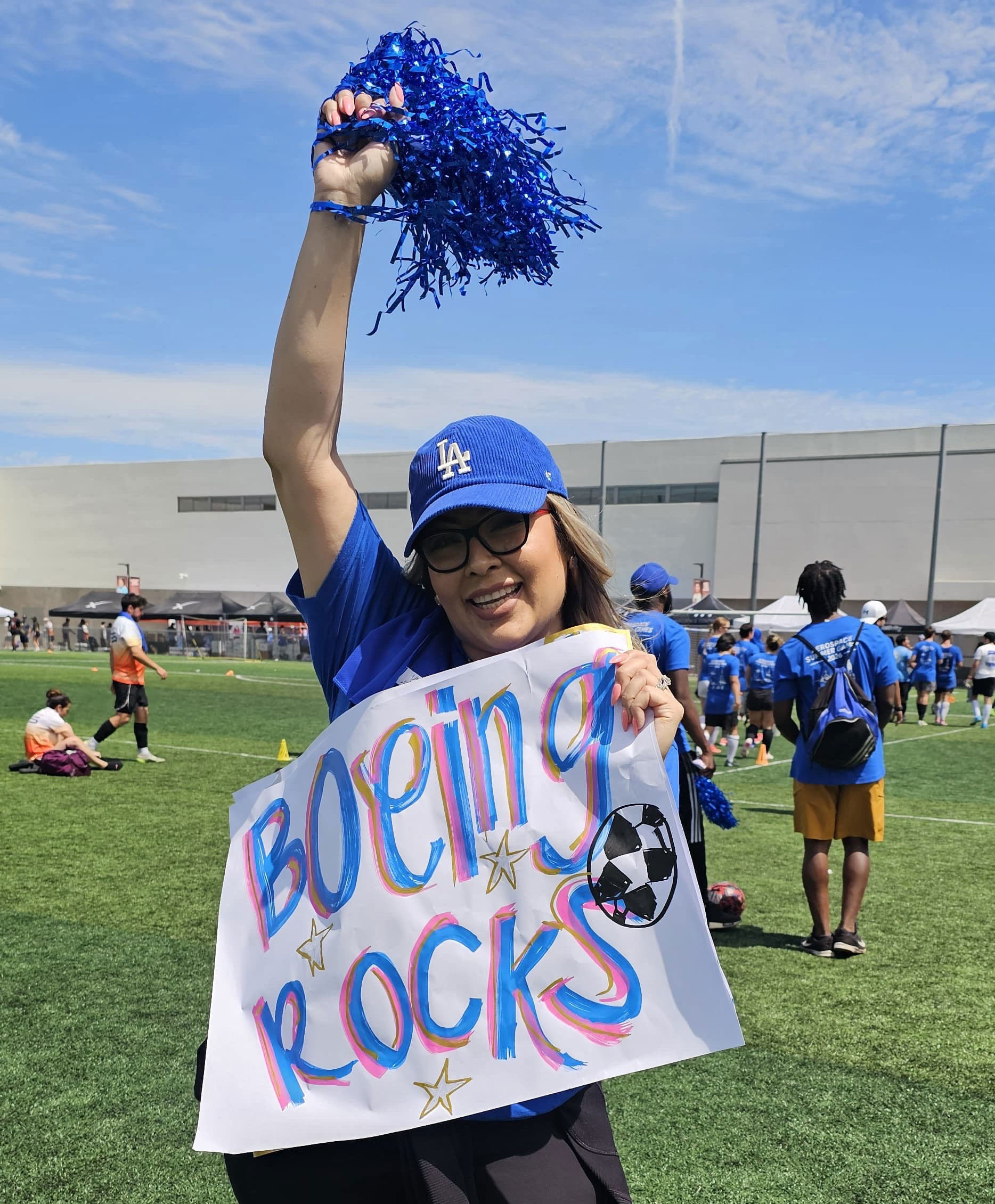 Woman smiling with a cheer pom pom in one hand and holding a "Boeing Rocks" poster with blue writing in the other.