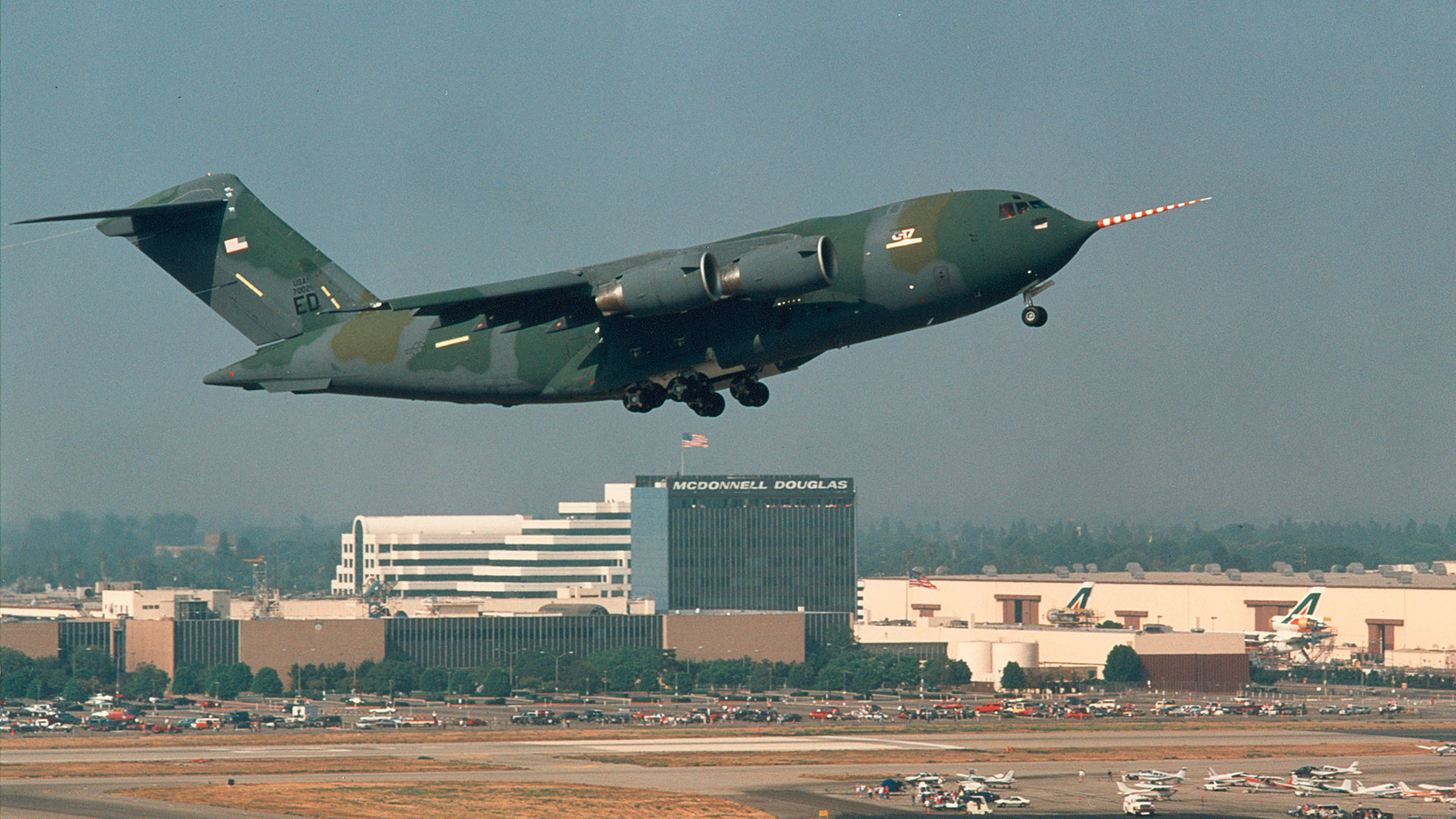 A large military cargo aircraft is flying low over an airport runway during landing approach, with its landing gear down and nose light visible. In the background are airport buildings, including a building labeled “McDonnell Douglas,” along with parked aircraft and runway areas under a hazy sky.