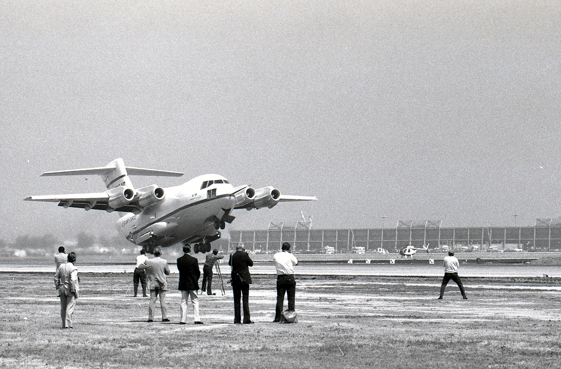 A black and white photo. A jet transport aircraft lifts off from a runway while several people stand nearby watching the takeoff.