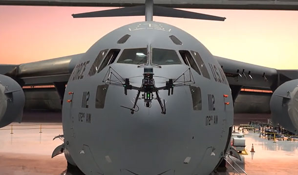 Front view of a large gray U.S. military cargo aircraft inside a hangar, with a drone mounted on its nose and the rear ramp open behind it.