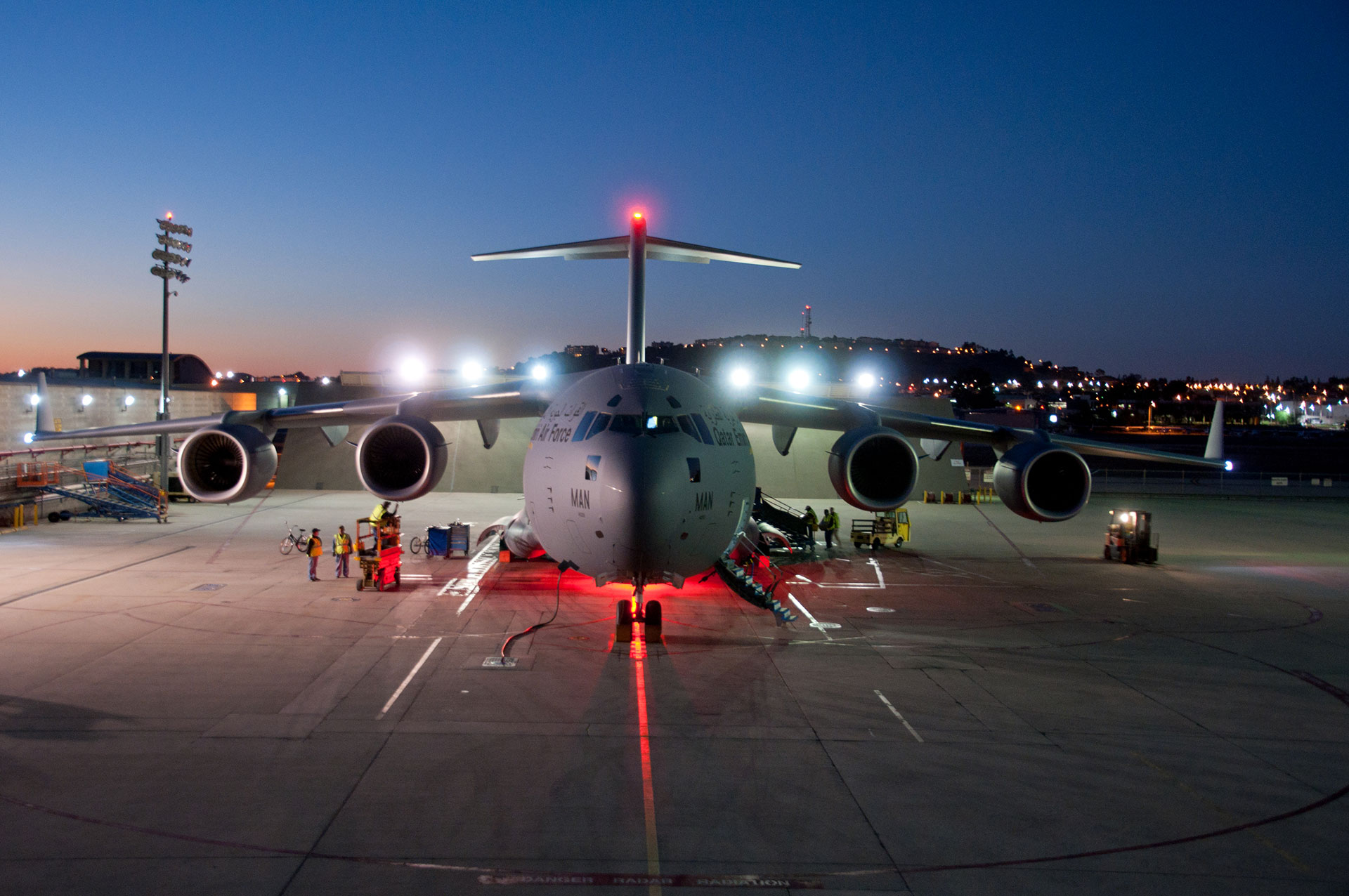 A large cargo aircraft is parked on a runway at dusk, viewed from the front with ground crew and service equipment around it under bright airport lights.