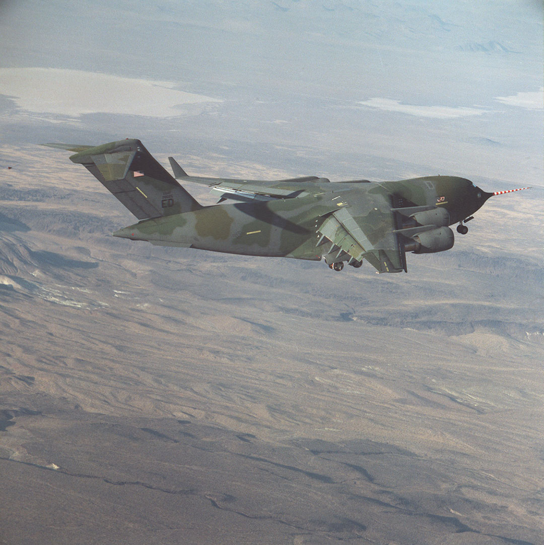 A camouflaged military cargo aircraft flies over a desert landscape, viewed from behind and slightly above, with its landing gear extended and a red-and-white striped probe extending from the nose.