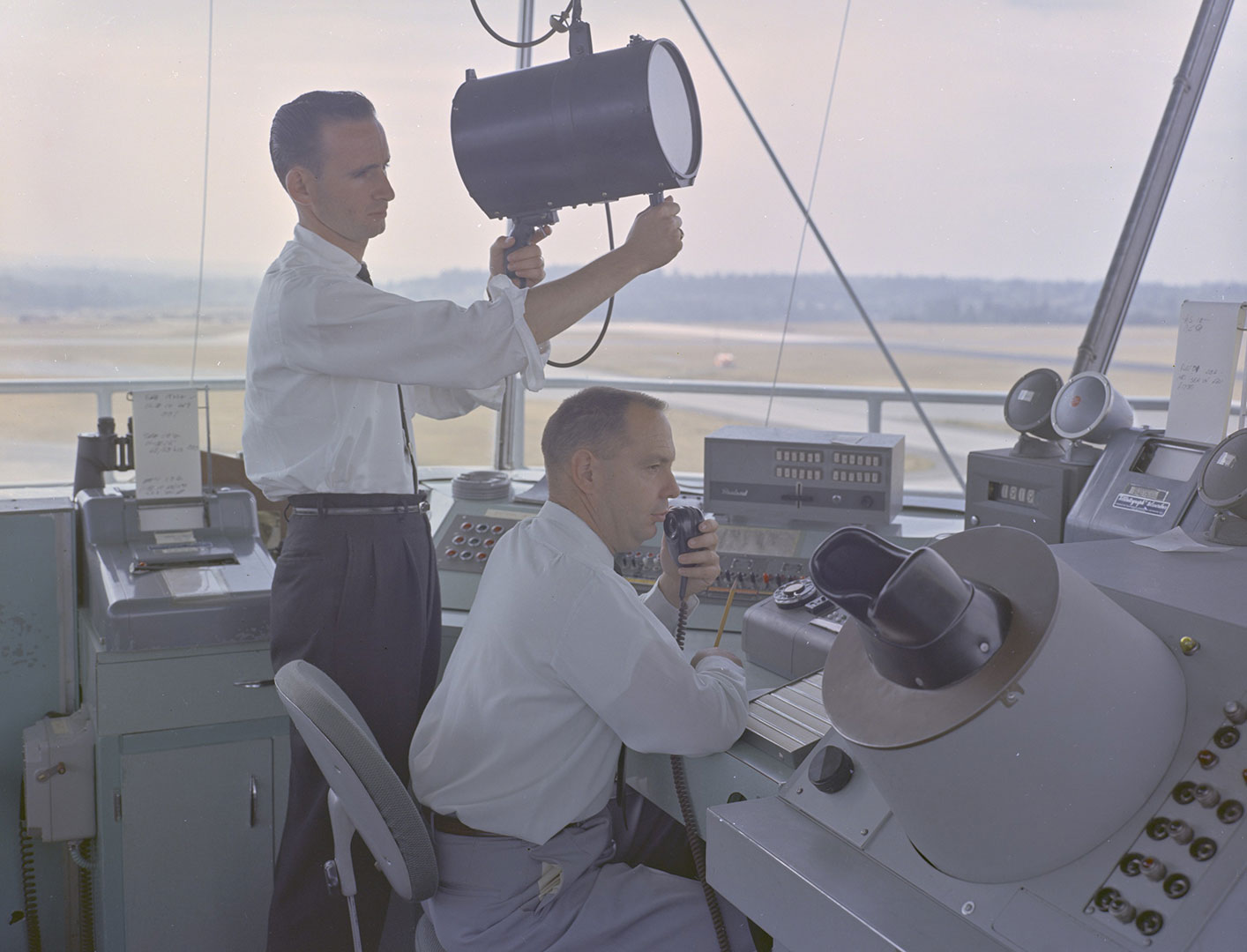 Color photo of two men in control tower with spot lights.