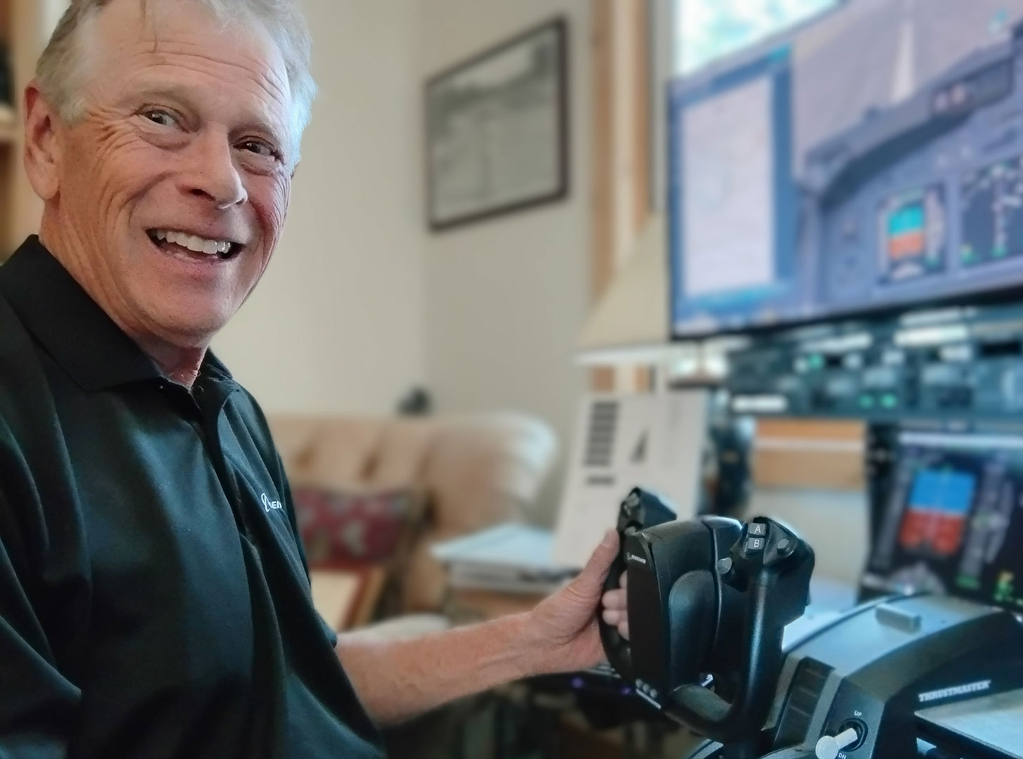 Bill Pieser smiles at the camera while sitting at the 737 flight simulator he built in his home in Colorado. Pieser is wearing a black Boeing polo shirt, and his left hand is touching the yoke of the simulator.