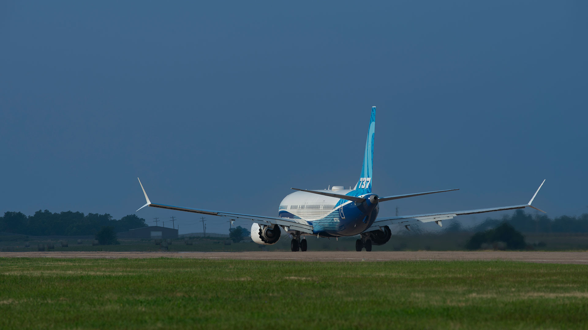 A Boeing 737 MAX-10 with Boeing livery performs a brake test on a runway. Green grass is in front of the plane and blue sky is behind it. The plane has traveled past the photographer, who is capturing the even from afar with a zoom lens.
