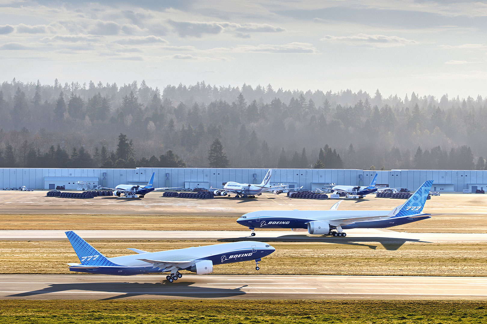 Boeing artist rendering of a 777 Freighter taking off in the foreground, with a 777-8 Freighter taxiing on the runway behind it. Several other freighters, including a 747-400 Freighter and additional 777 Freighters, are parked near airport buildings in the background. Trees and a hazy sky surround the airport.