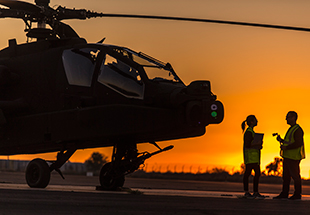  Silhouette of a man and woman talking next to an Apache helicopter. at sun set on tarmac. U.S. Army Services 