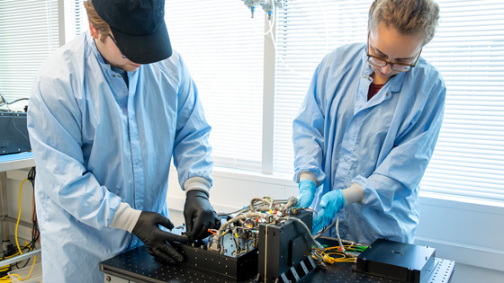 Two technicians in lab coats working on electronic equipment in a cleanroom environment.