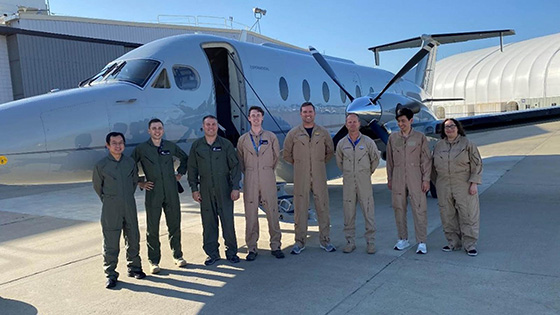 A group of eight technicians in flight suits standing in front of a small aircraft on a sunny day.