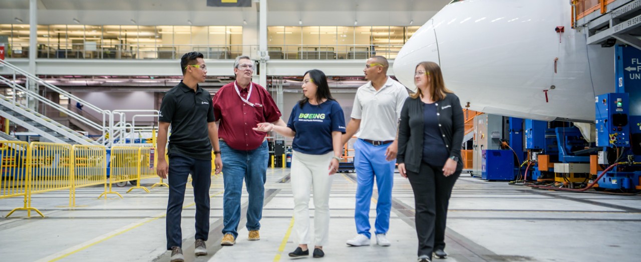 Five Boeing employees of varying ages, genders and races walk across a factory floor, talking with each other. Behind them is the nose of a 787.
