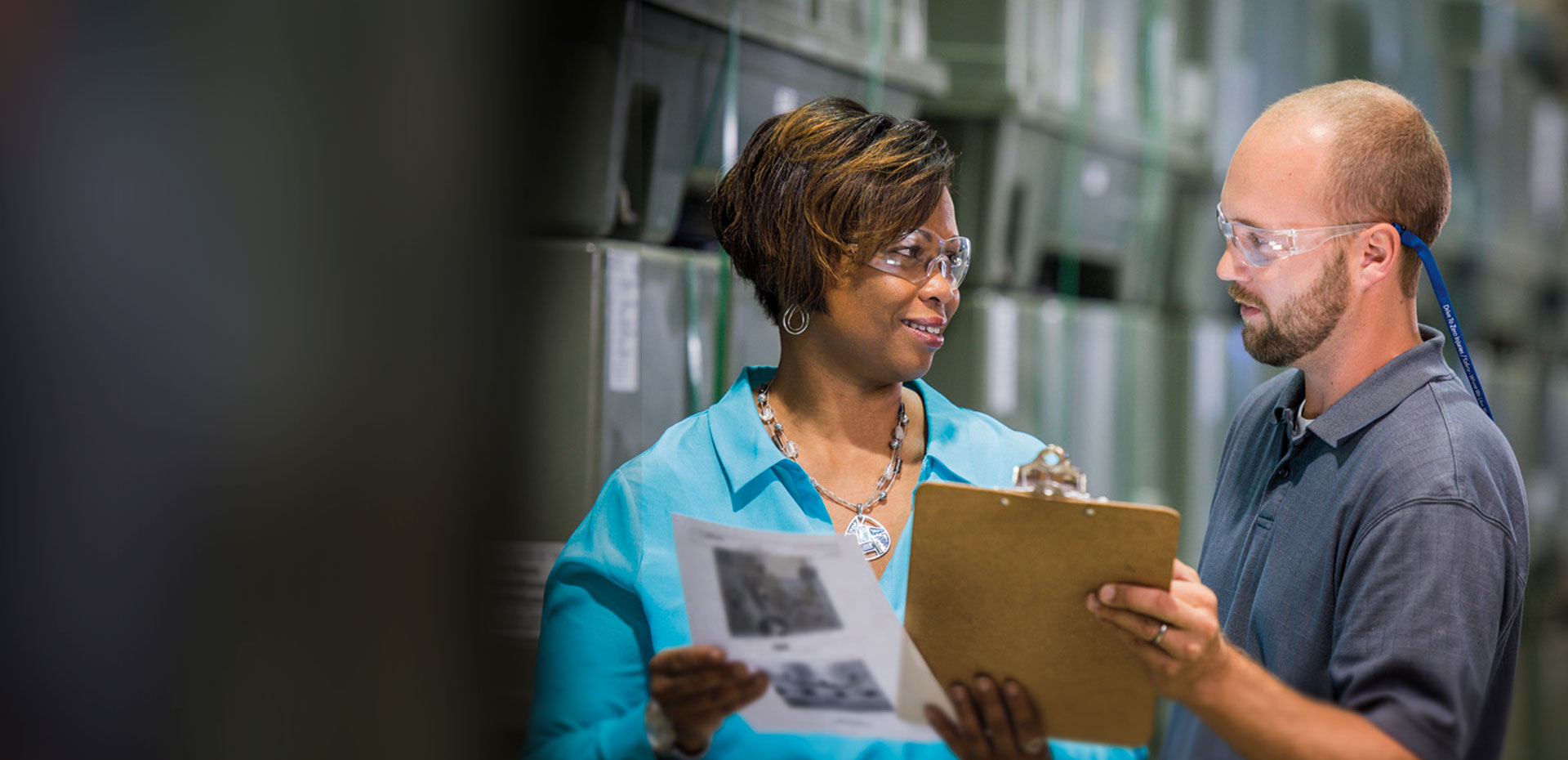 Two people looking at a clipboard in front of a set of shelves 